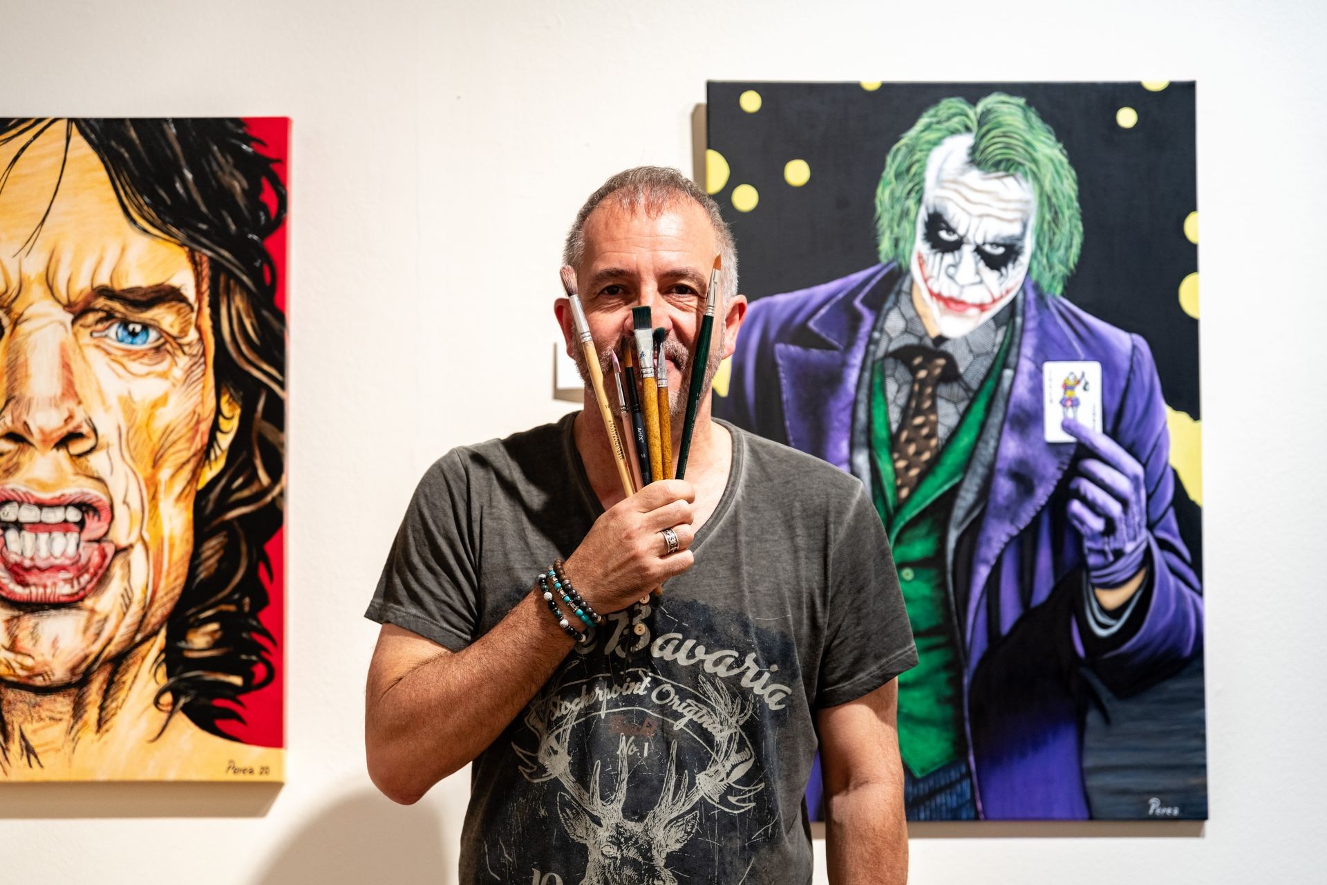 Artist holding paintbrushes stands between two portrait paintings on a wall.