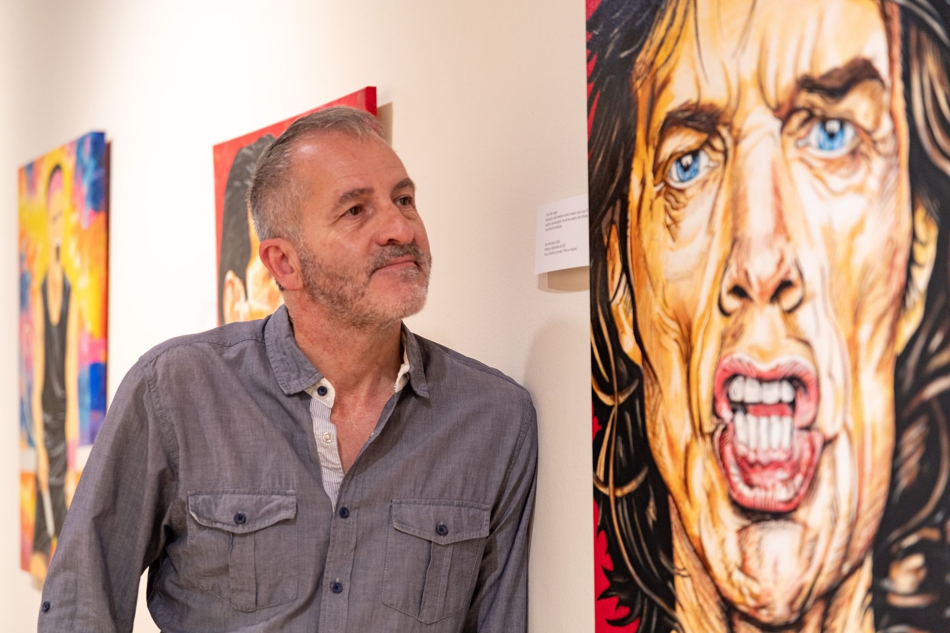 Person in a denim shirt standing in an art gallery looking at colorful paintings on the wall.