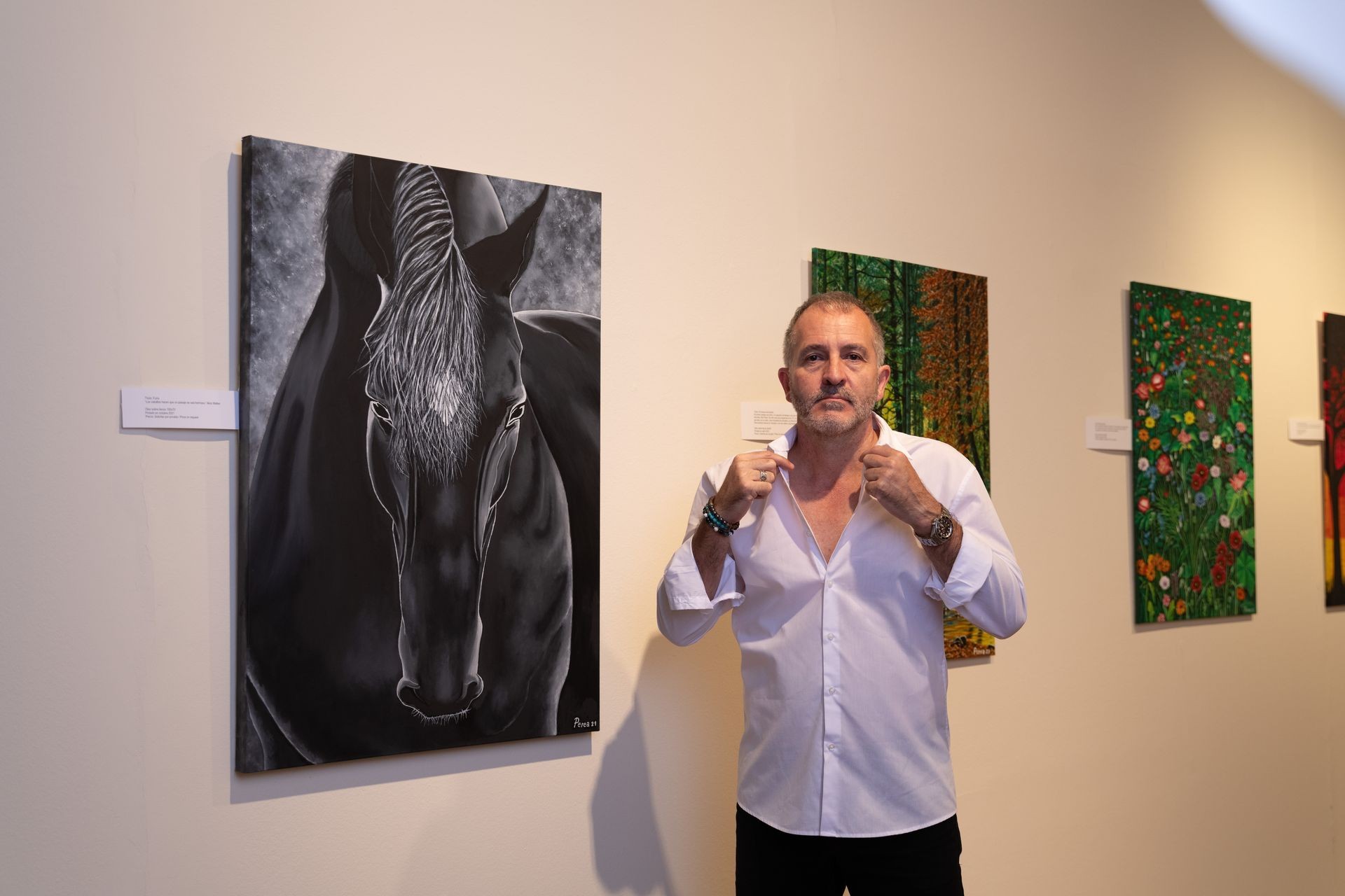 Man standing beside a black horse painting on gallery wall, adjusting his shirt collar.